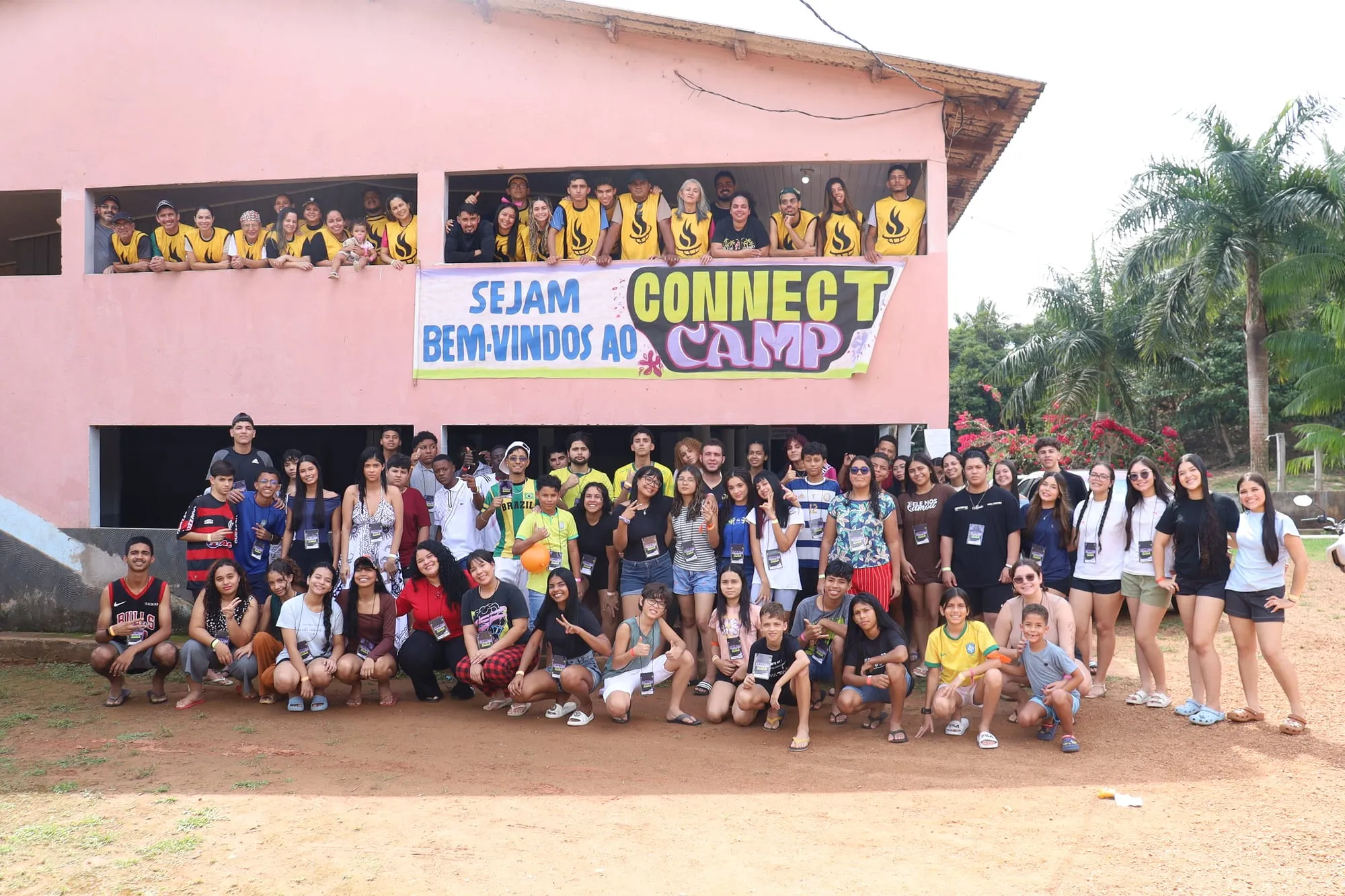 Foto do grupo completo do acampamento reunido em frente ao prédio, sob a faixa 'Sejam bem-vindos ao Connect Camp'