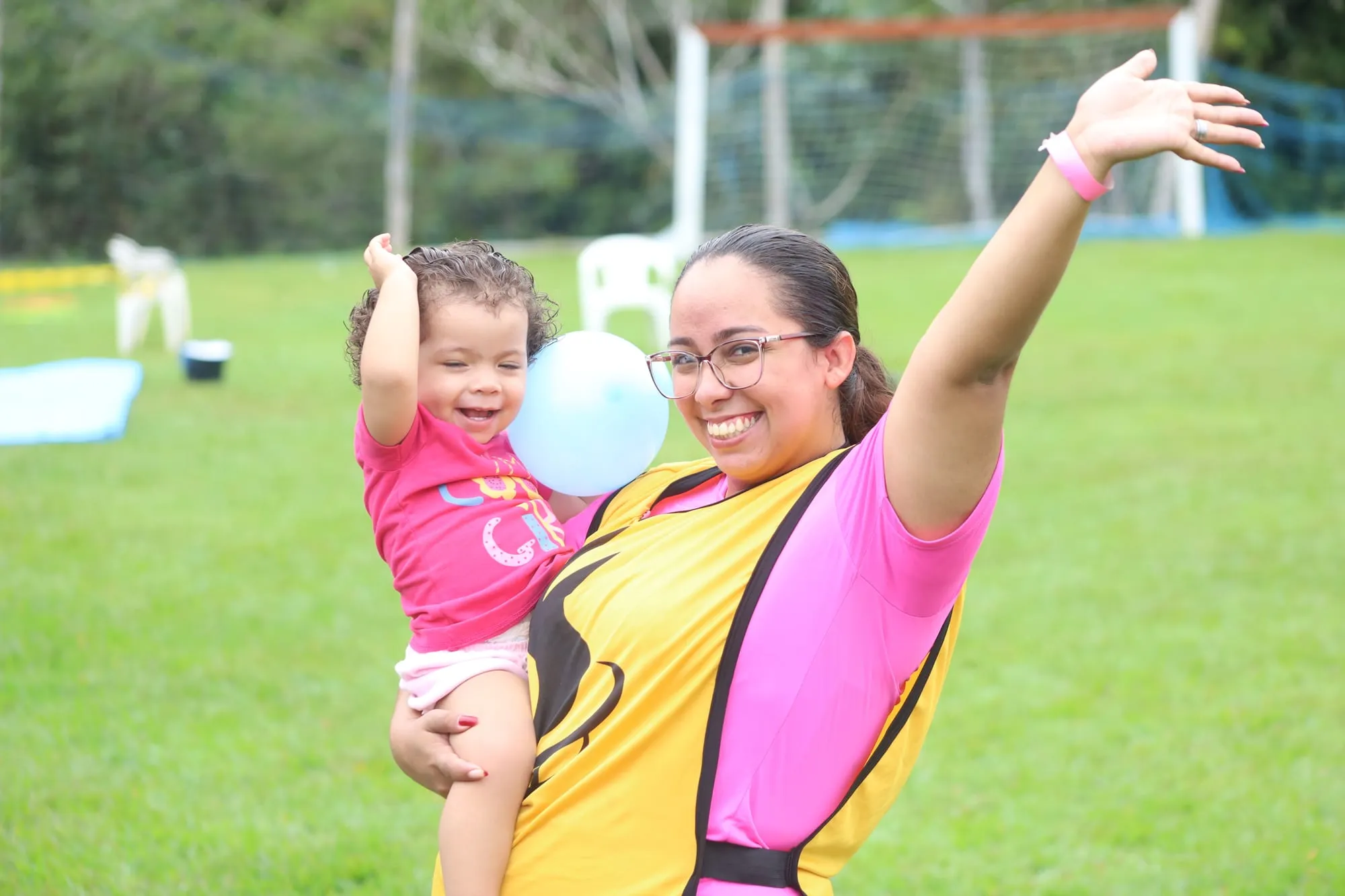 Voluntária de colete sorrindo com o braço erguido enquanto segura no colo uma criança pequena com um balão azul, no gramado