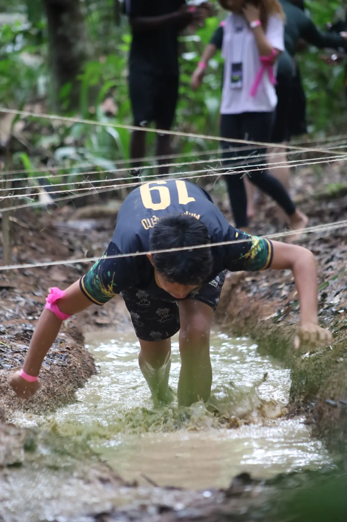 Jovem de camiseta número 19 atravessando de quatro a poça de lama do circuito de obstáculos, passando por baixo das cordas