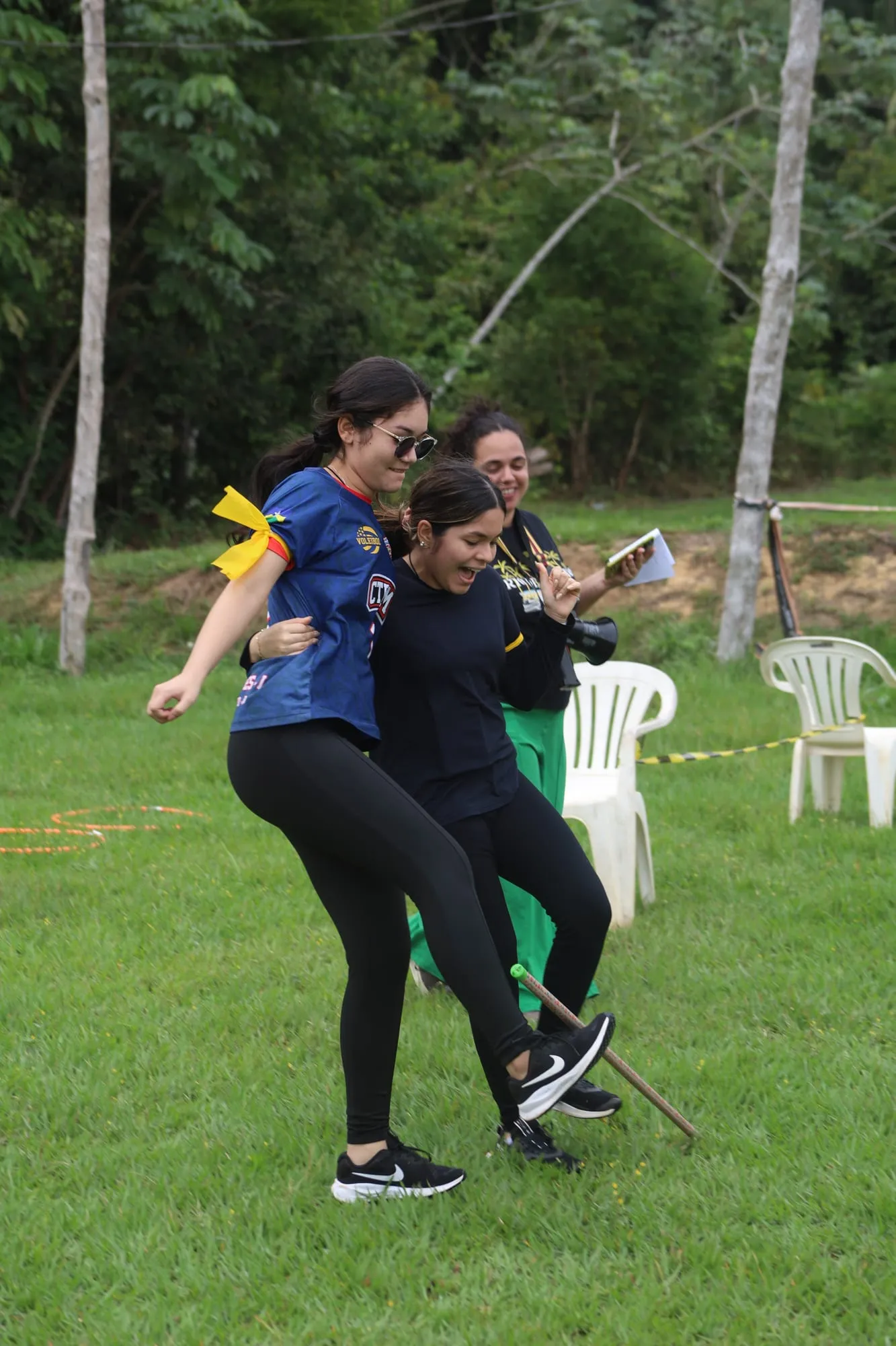 Duas jovens rindo durante uma brincadeira em equipe no gramado, com uma líder segurando um megafone ao fundo