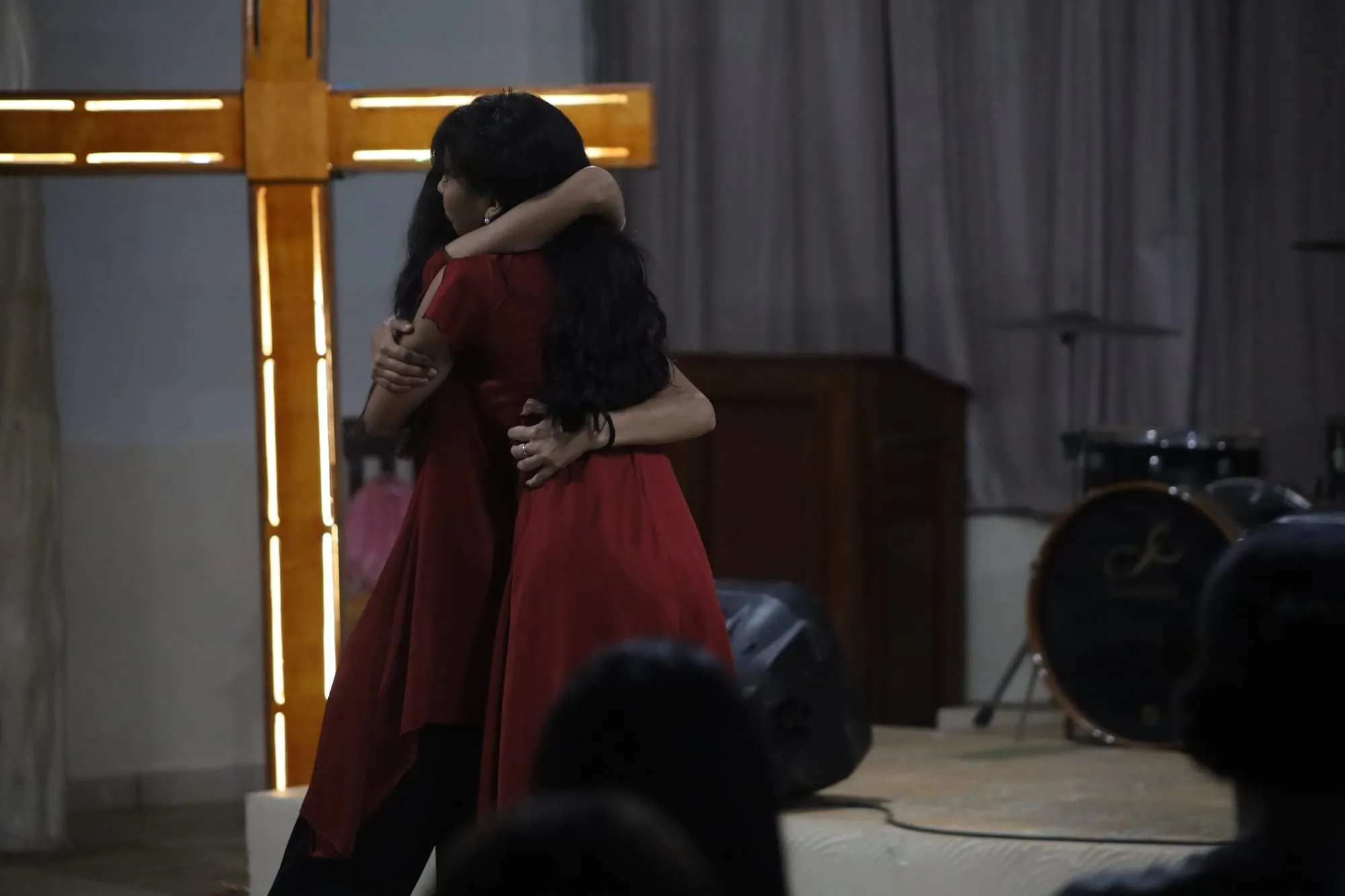 Duas jovens de vestido vermelho se abraçando diante de uma cruz de madeira iluminada no palco