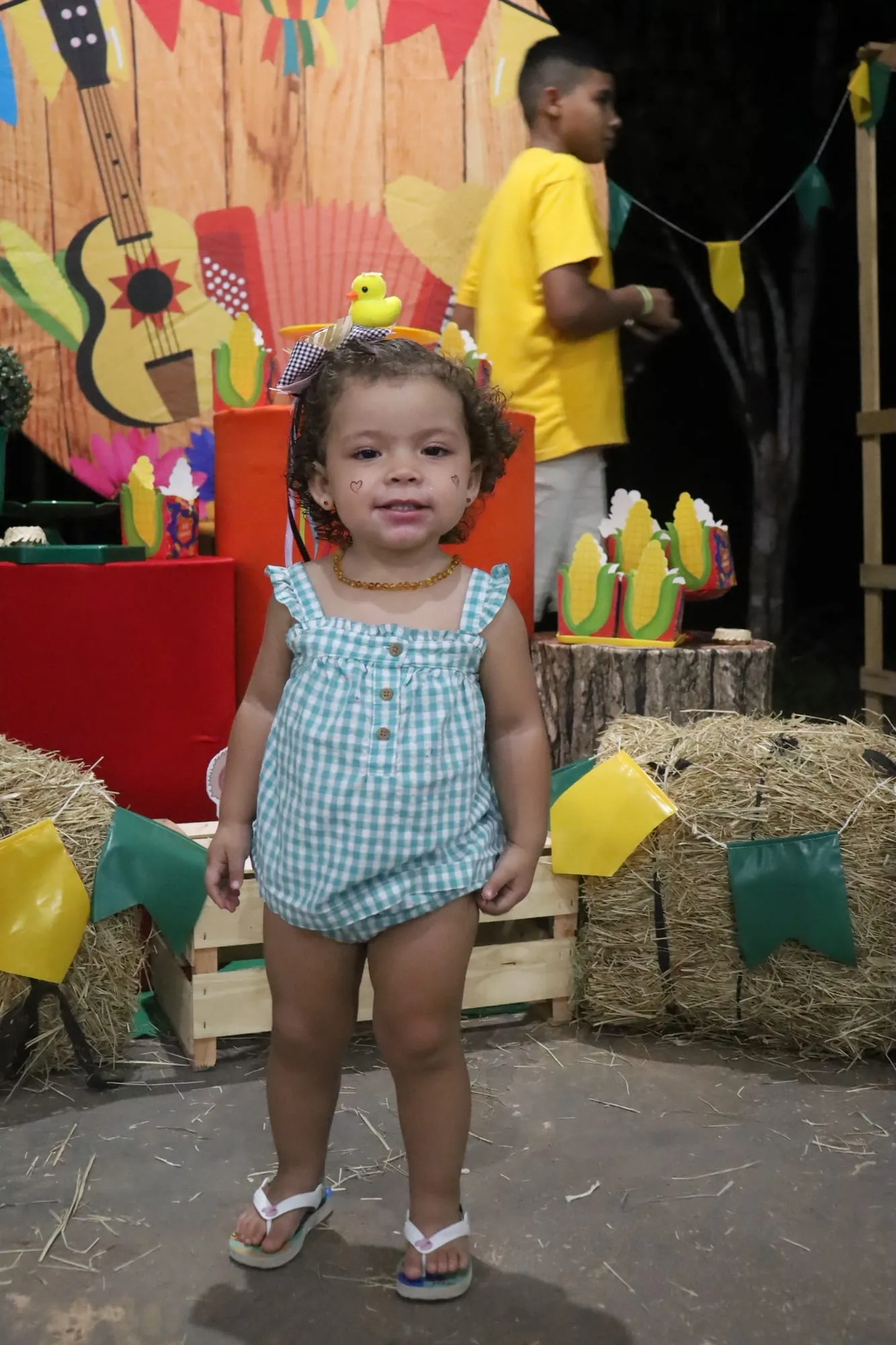 Menininha de macacão xadrez sorrindo diante da decoração de festa junina