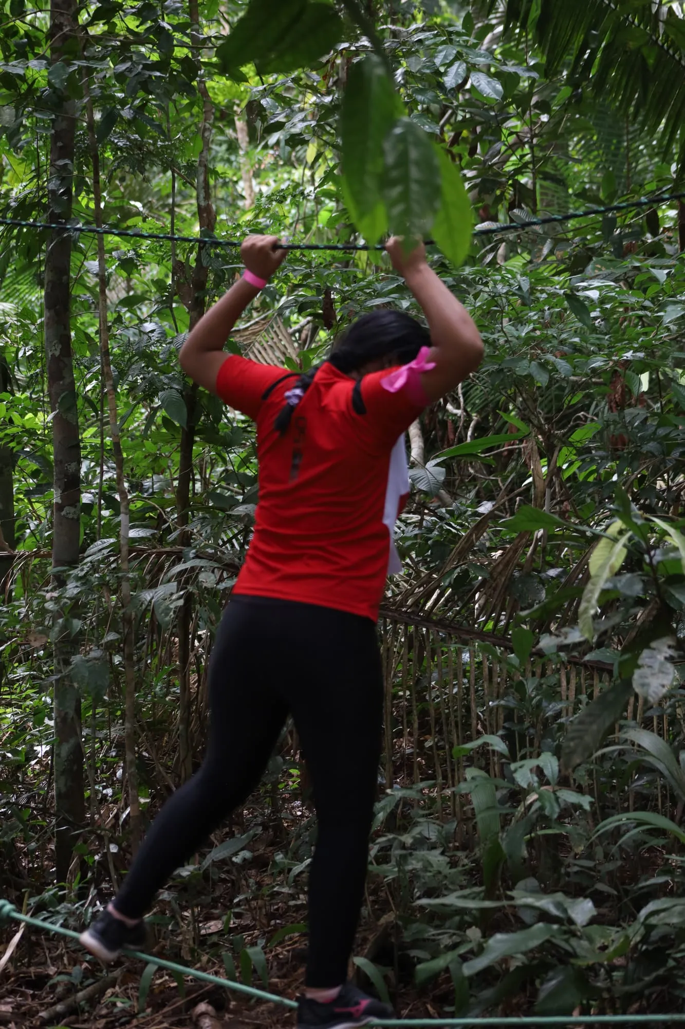 Jovem de camiseta vermelha vencendo a travessia na corda do circuito de obstáculos em meio à mata