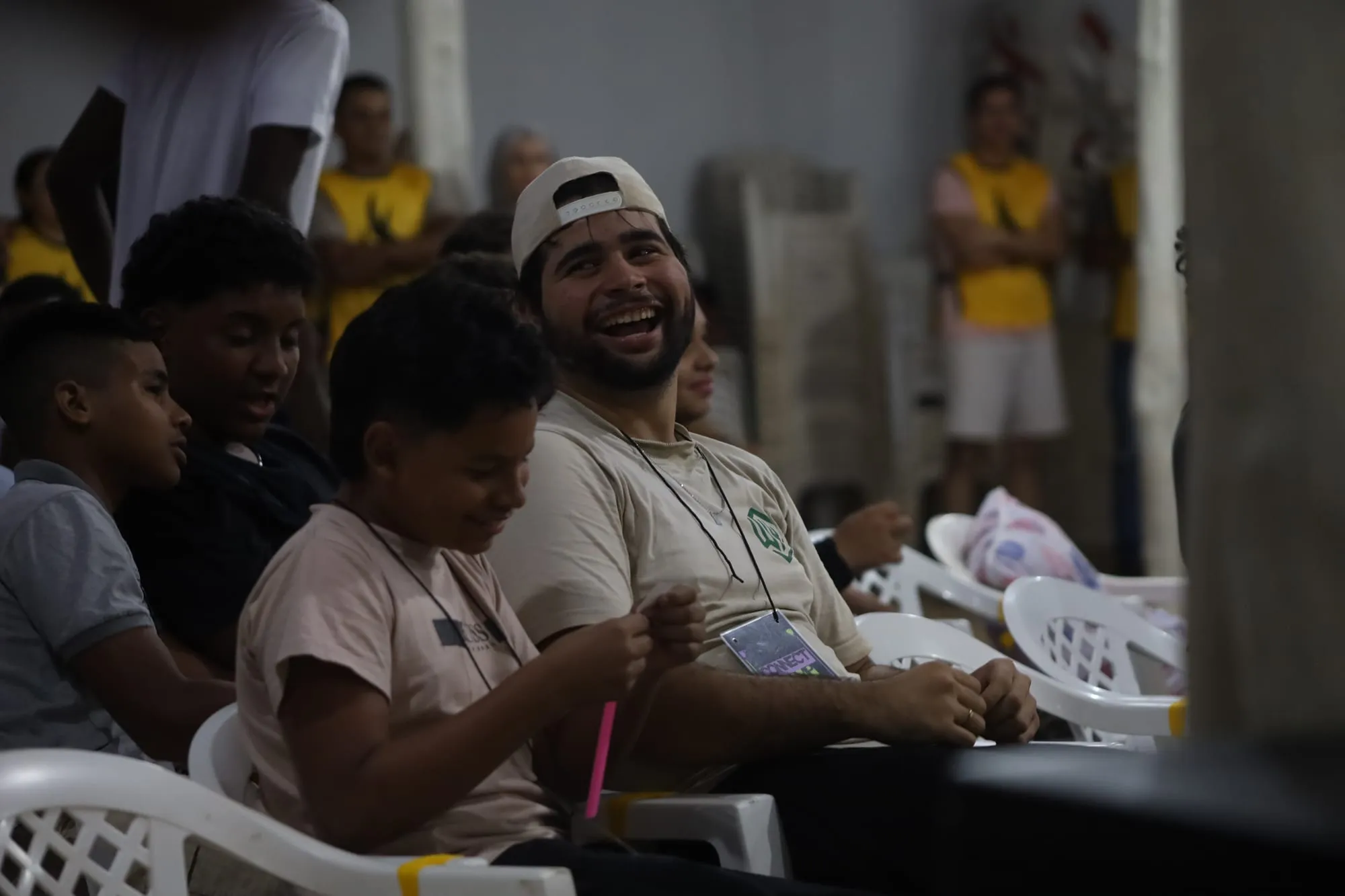 Rapaz de boné rindo animado, sentado no meio da garotada durante uma atividade