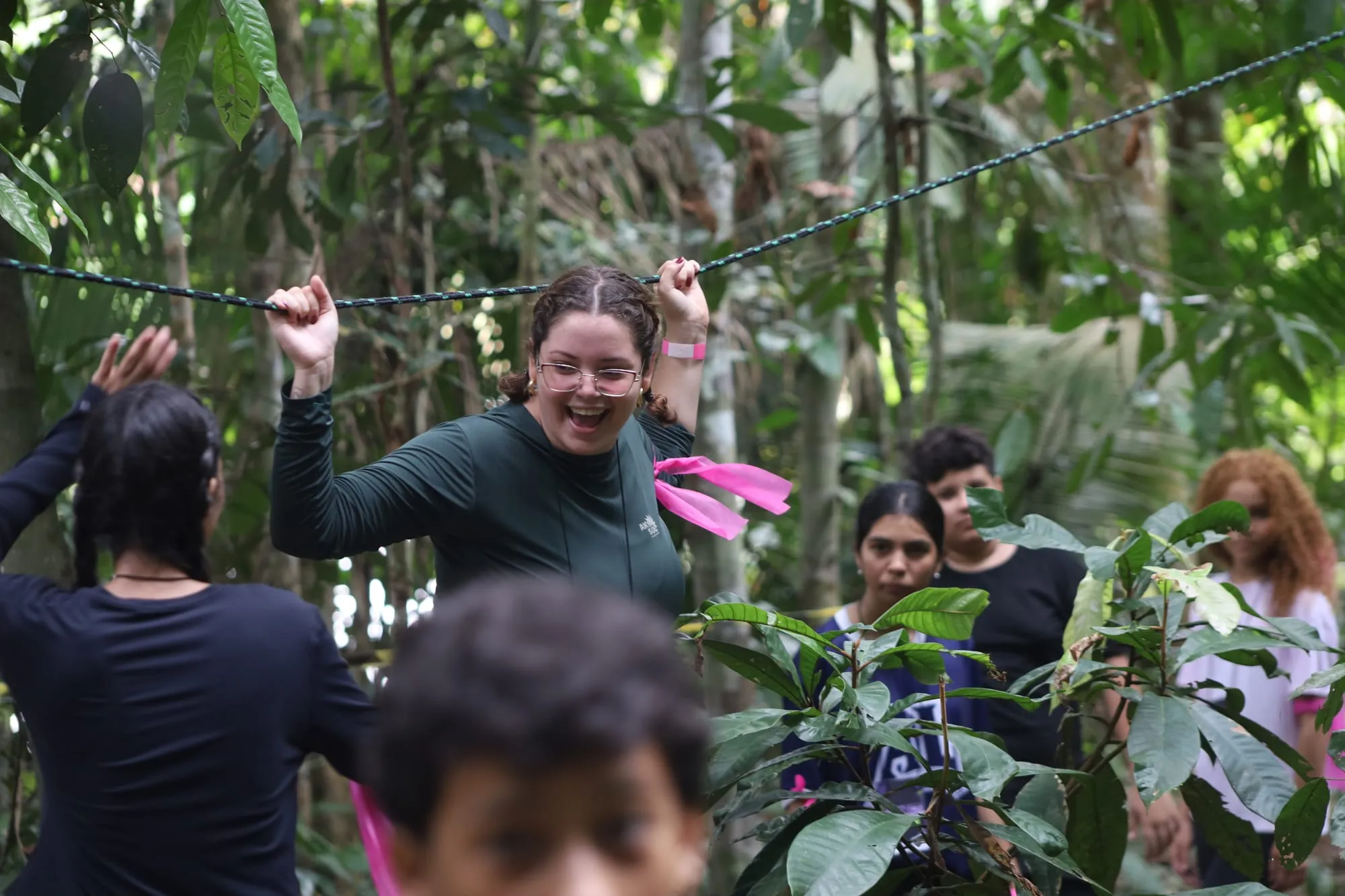 Mulher rindo enquanto se equilibra na corda do circuito de obstáculos na mata, cercada pelo grupo