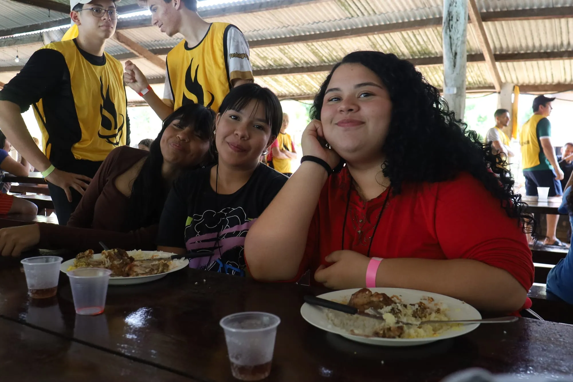 Três jovens sorrindo juntas à mesa durante a refeição, com voluntários de colete ao fundo