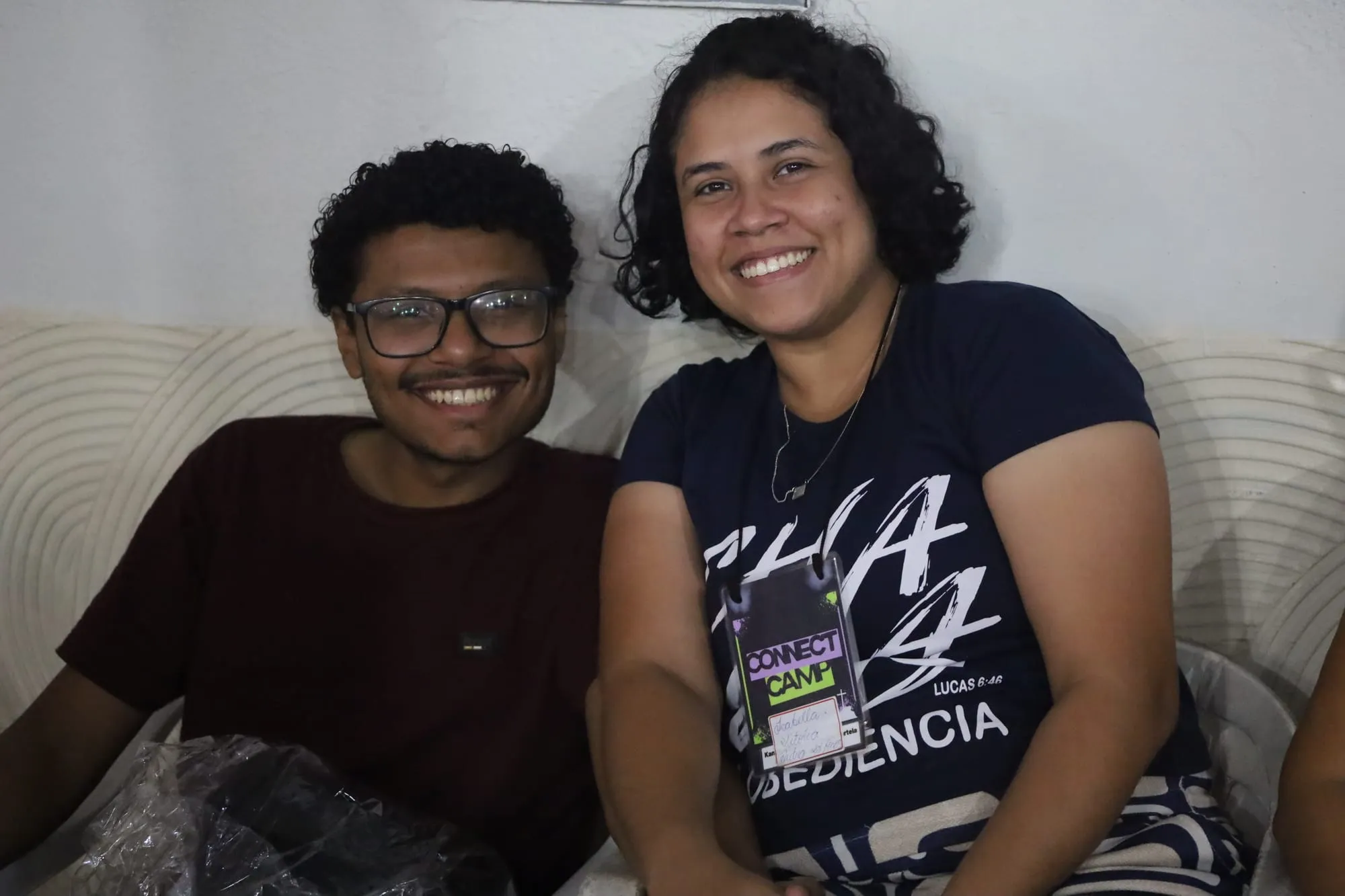 Dois amigos sorrindo lado a lado no sofá, um de óculos e camiseta vinho e a outra com o crachá do Connect Camp